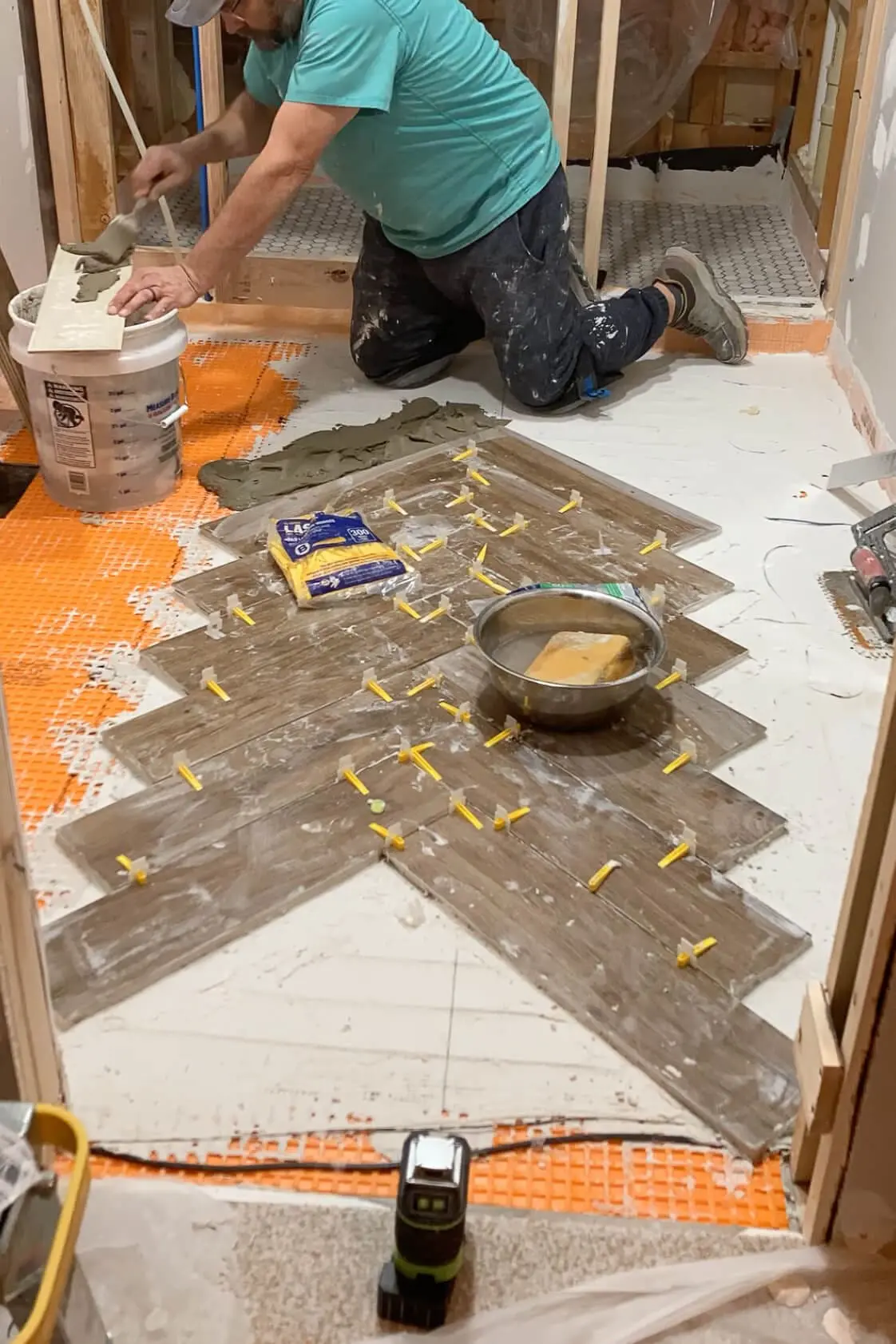 A worker applying mortar to the back of a tile while completing a herringbone wood-look tile pattern on a bathroom floor with a Schluter-DITRA underlayment visible.